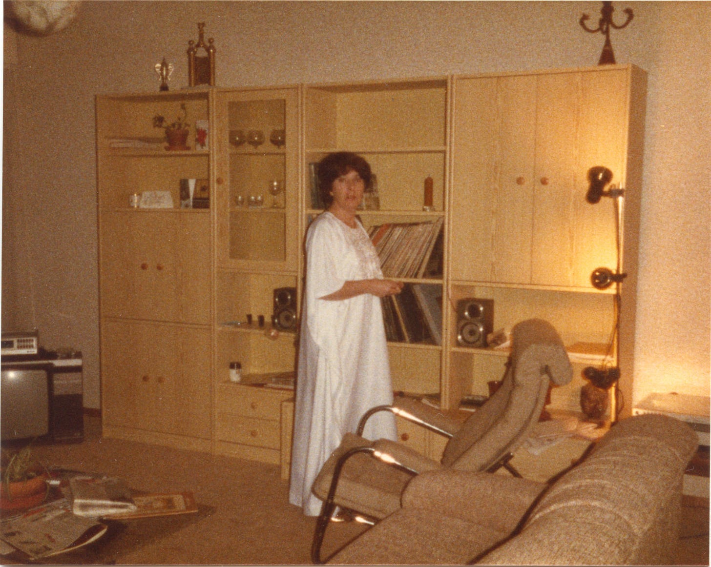 A woman dressed in a floor length white satin kaftan in front of a large display cabinet in a flat