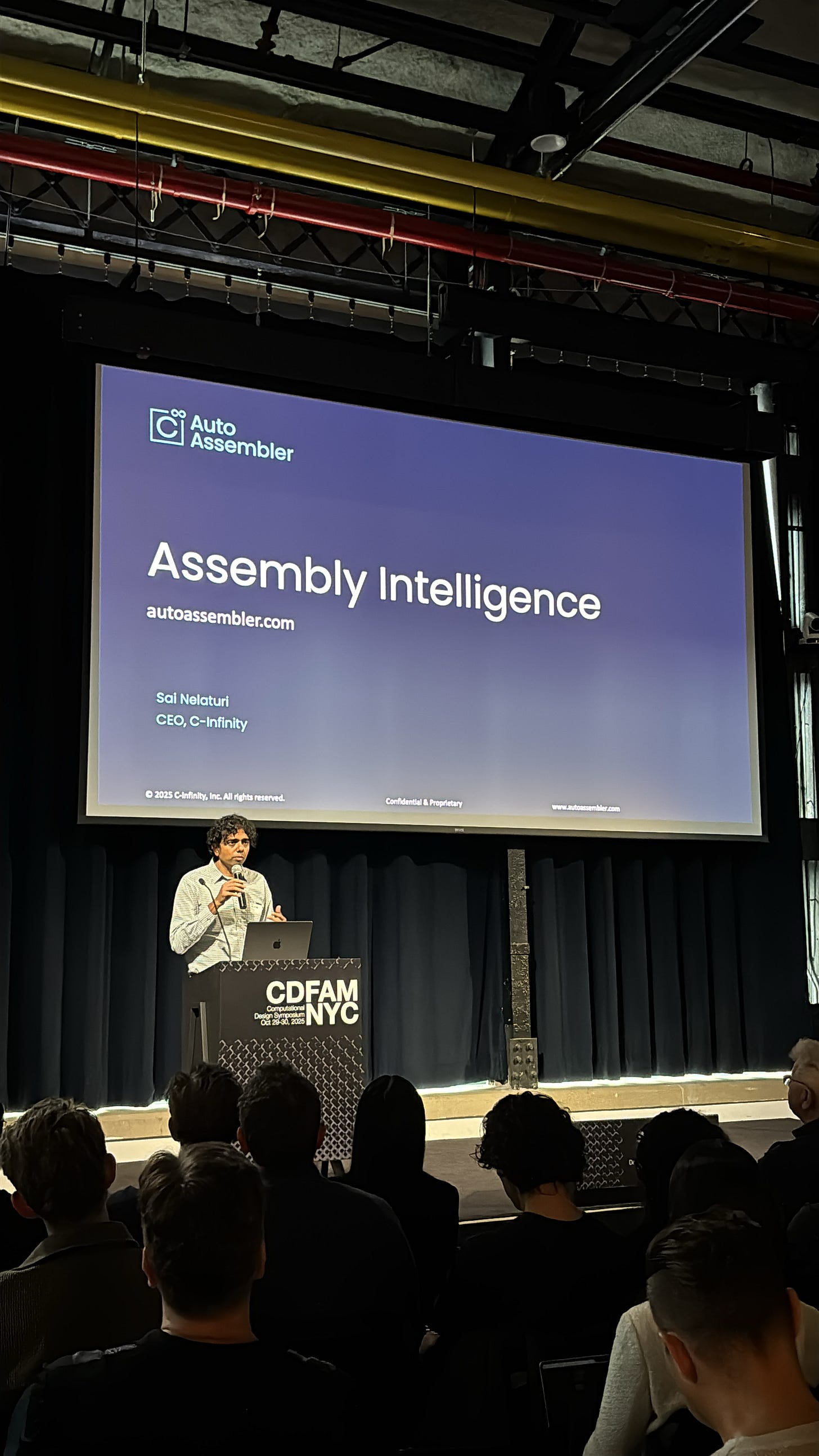 A speaker presenting on stage at an event, with a large screen behind showing the title 'Assembly Intelligence' and the presenter's name. The audience is visible in the foreground.