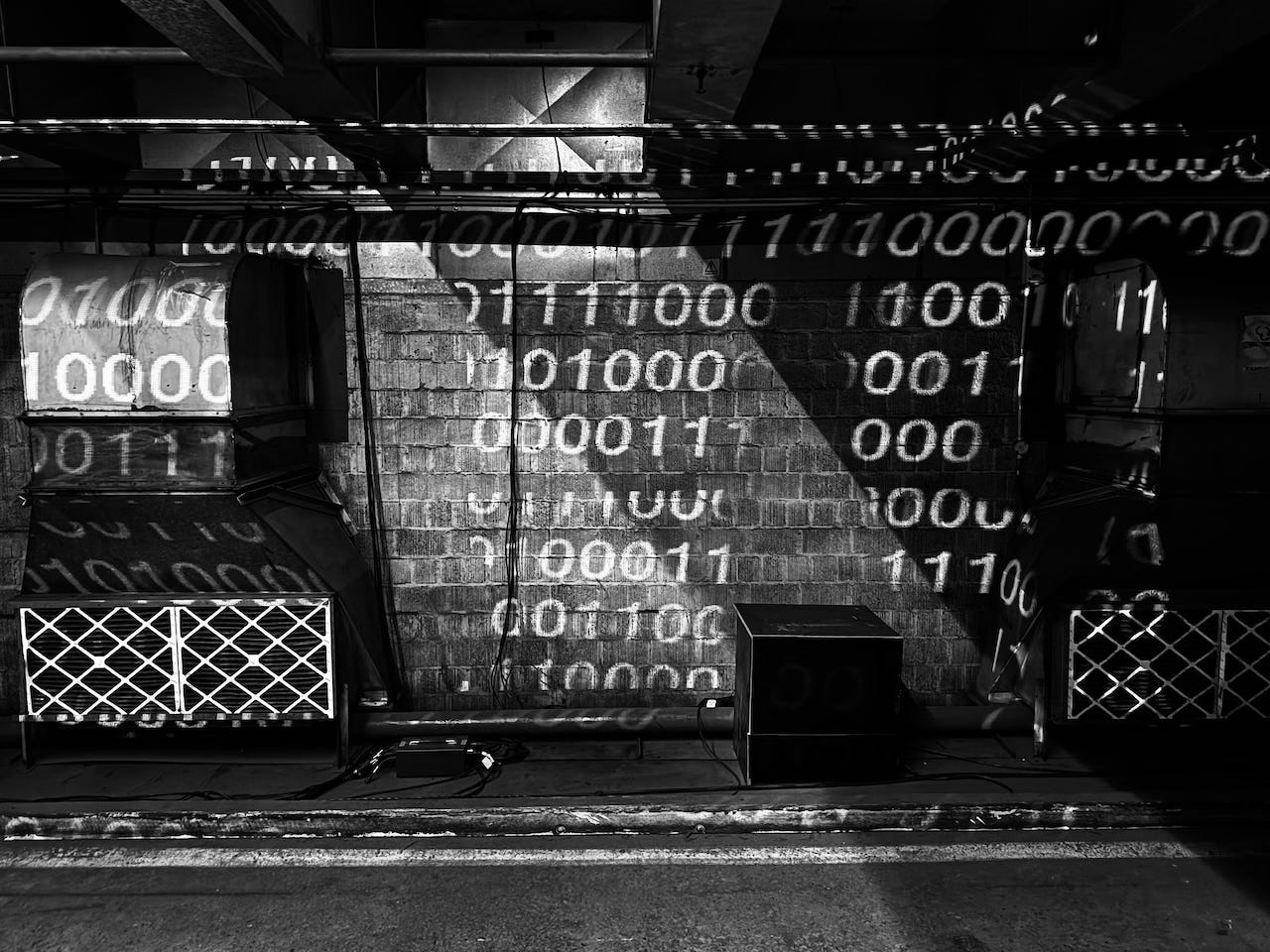 GPT-4o: A black-and-white photo showing a gritty industrial setting with a brick wall, ventilation ducts, and metallic grates. Projected onto the wall and surrounding surfaces are sequences of large binary numbers (“1s” and “0s”). These numbers create patterns of light and shadow, suggesting a digital or cyber theme. In the foreground, a metal grate structure is visible, along with cables and a box-like object on the ground. The image conveys a fusion of technology and an industrial environment, evoking themes of data, systems, and digital complexity.