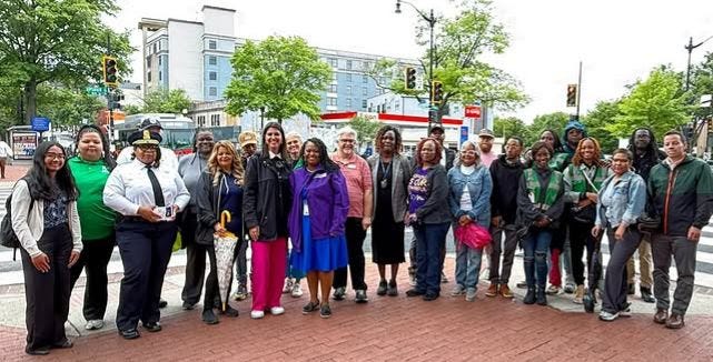 Photo of CM Brooke Pinto (in pink pants) and CM Janeese Lewis George (in purple jacket) in front of a semi-circle of community members, MPD, Safe Passage Team members.