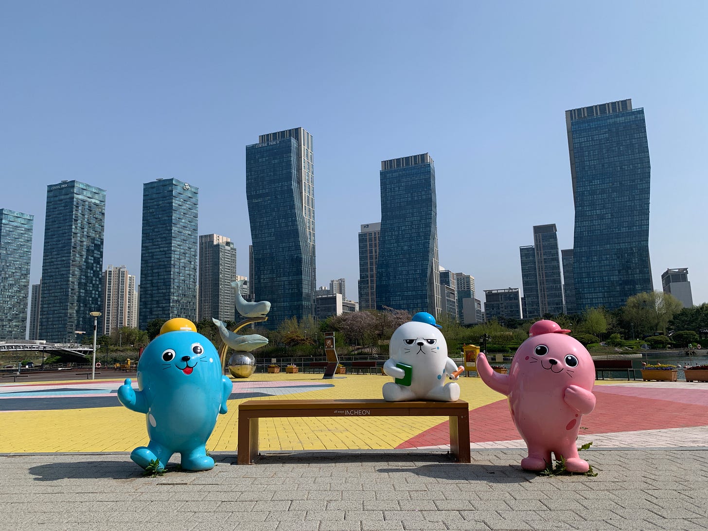 A photo of a street scene in Incheon, South Korea. In the foreground are three statues of cute animated animals around a park bench and in the background are rows of gleaming skyscrapers