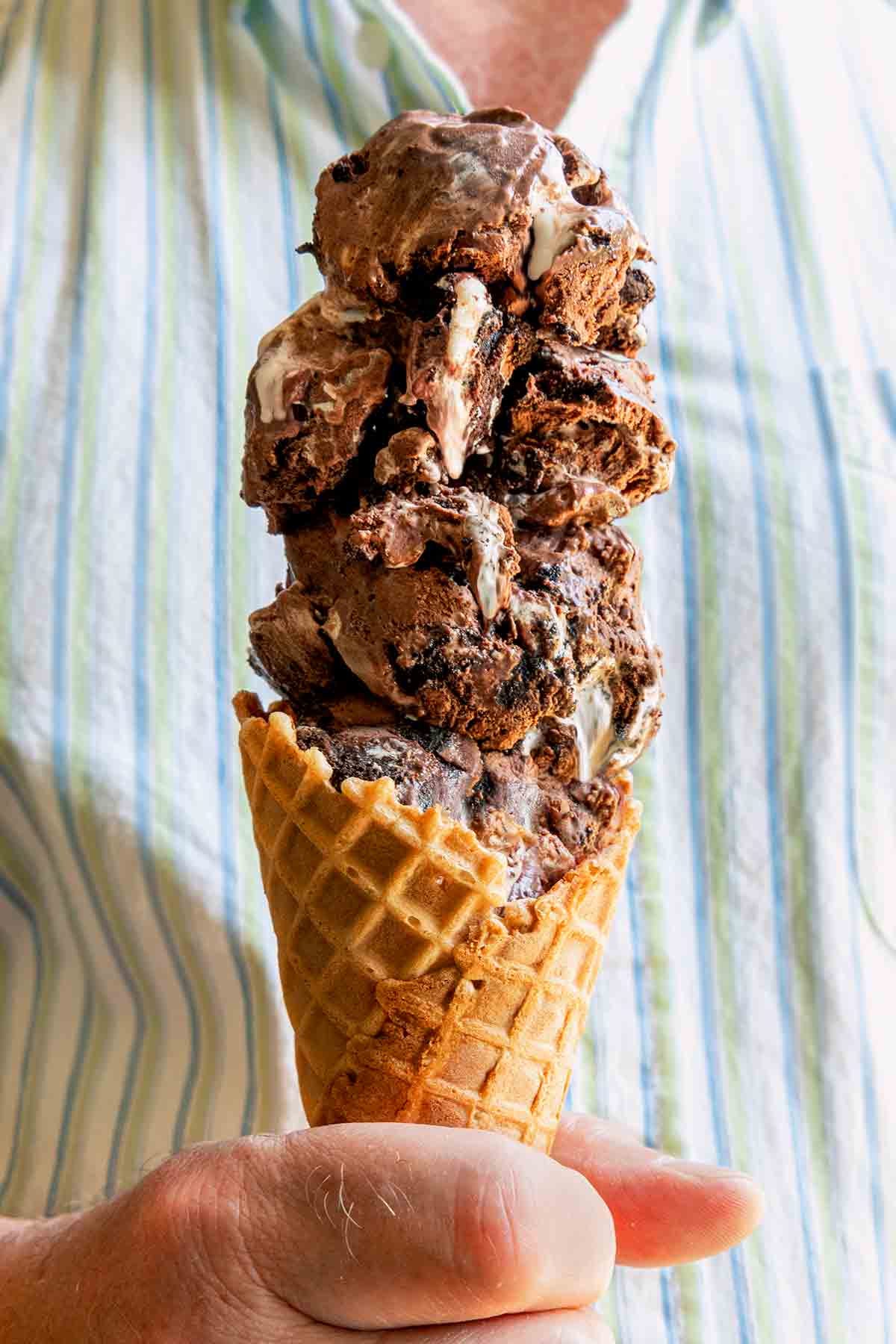 A close-up of a man's hand holding an ice cream cone with four scoops of Chocolate Oreo Ice Cream.