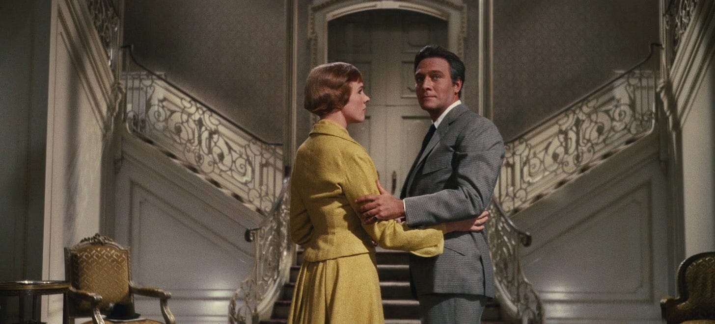 A screenshot from Sound of Music, depicting Maria--a woman with short red hair wearing a yellow dress--looking with concern at Captain Von Trapp--a dark-haired man in a grey suit, looking sadly at the foyer of their home.