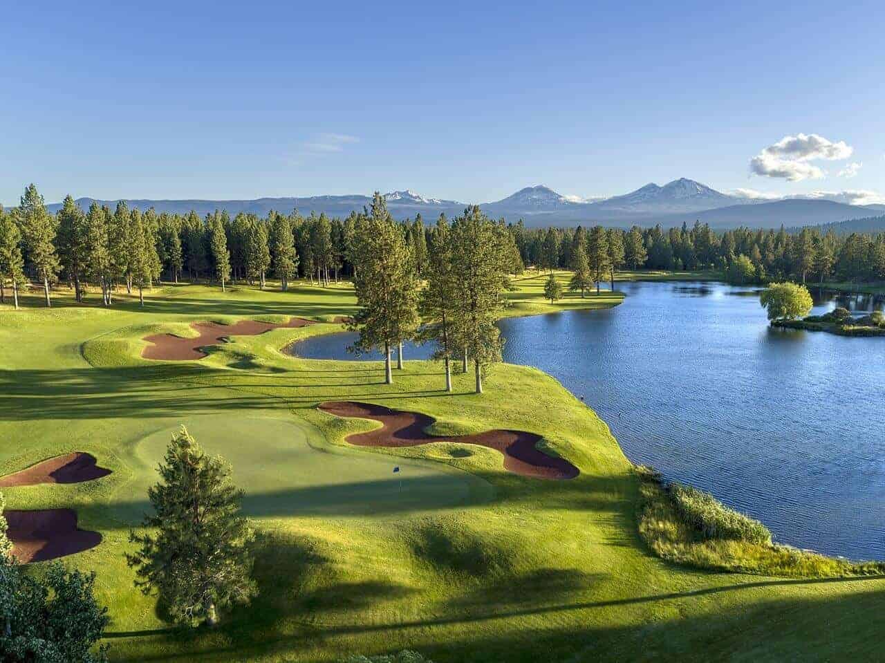 Aerial view of a lush green golf course with red sand bunkers, surrounded by pine trees and a lake, with snow-capped mountains in the background. Aerial view of a lush green golf course with red sand bunkers, surrounded by pine trees and a lake, with snow-capped mountains in the background.
