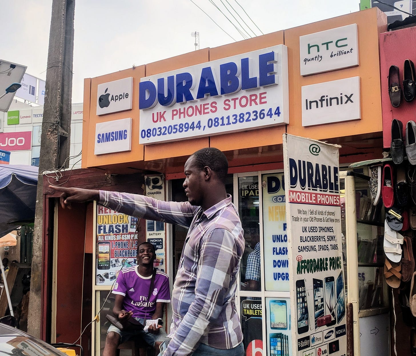 One man sits in front of a phone store with a sign that reads "Durable UK Phones Store." Another man beside him points in the distance.