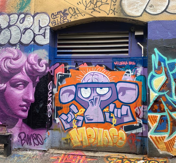 Two street murals, one featuring a realistic purple painting of Michelangelo's David's head next to a painting by another artist of a purple monkey head. The other image is of Michelangelo's Laocoön in pink, painted in a similarly realistic style.