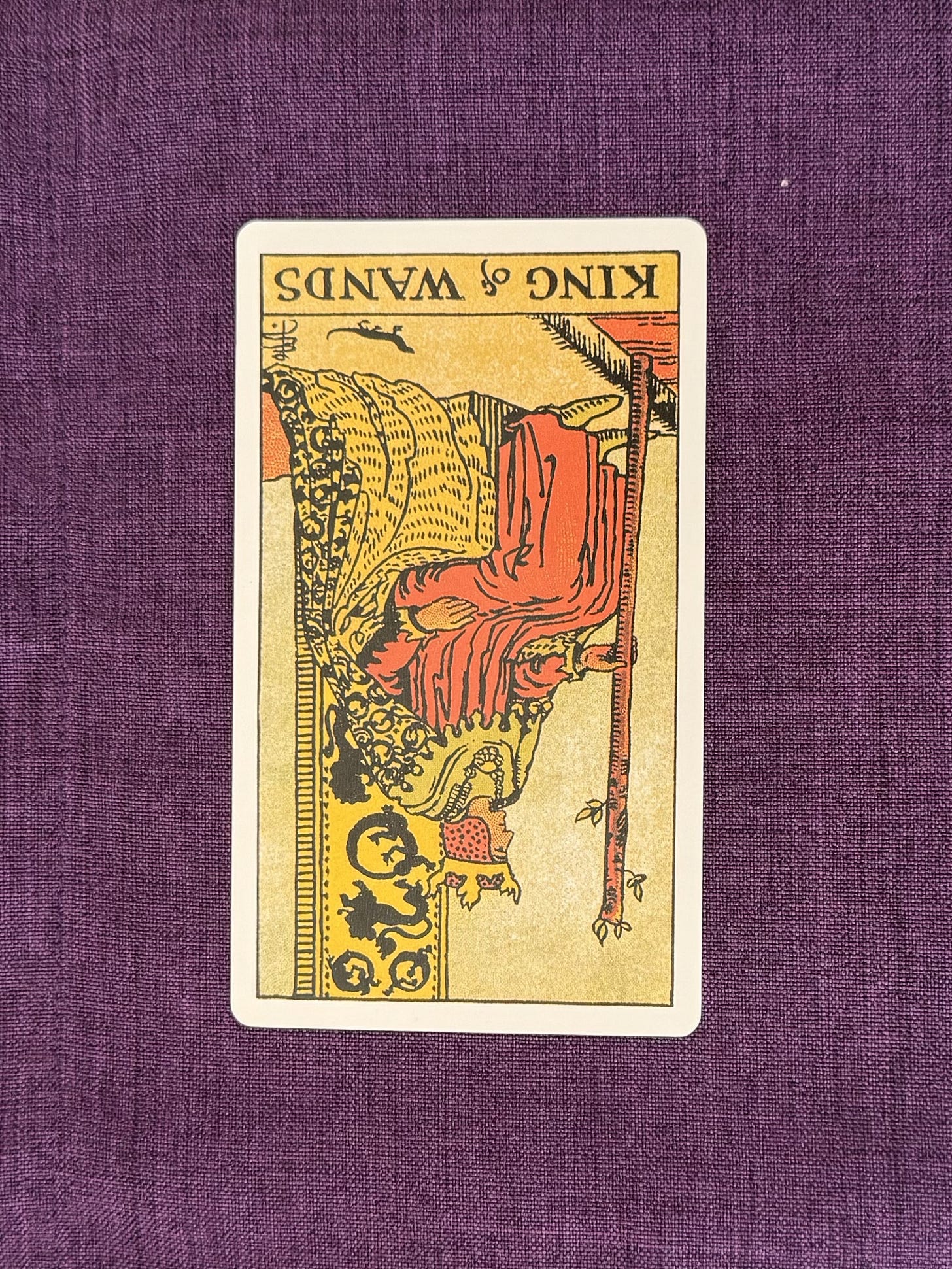Tarot card on a purple linen cloth shows The King of Wands depicts a man who has in his hands a blossoming wand. His throne, as well as his cape, are decorated with the salamander biting its tail and the lion. The robe is bright and orange resembling a flame, while the crown is shaped like a tongue of fire.