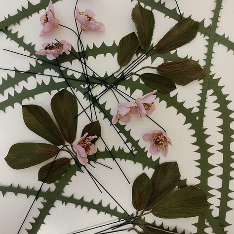 Image 1 is a cup full of mauve flowers, image 2 a collection of yellow pea- style flowers and deep green spikes, Image 3 is a series of cutout green stems with thorns with some dark green leaves and pink flowers. All are made of paper and depict works in progress