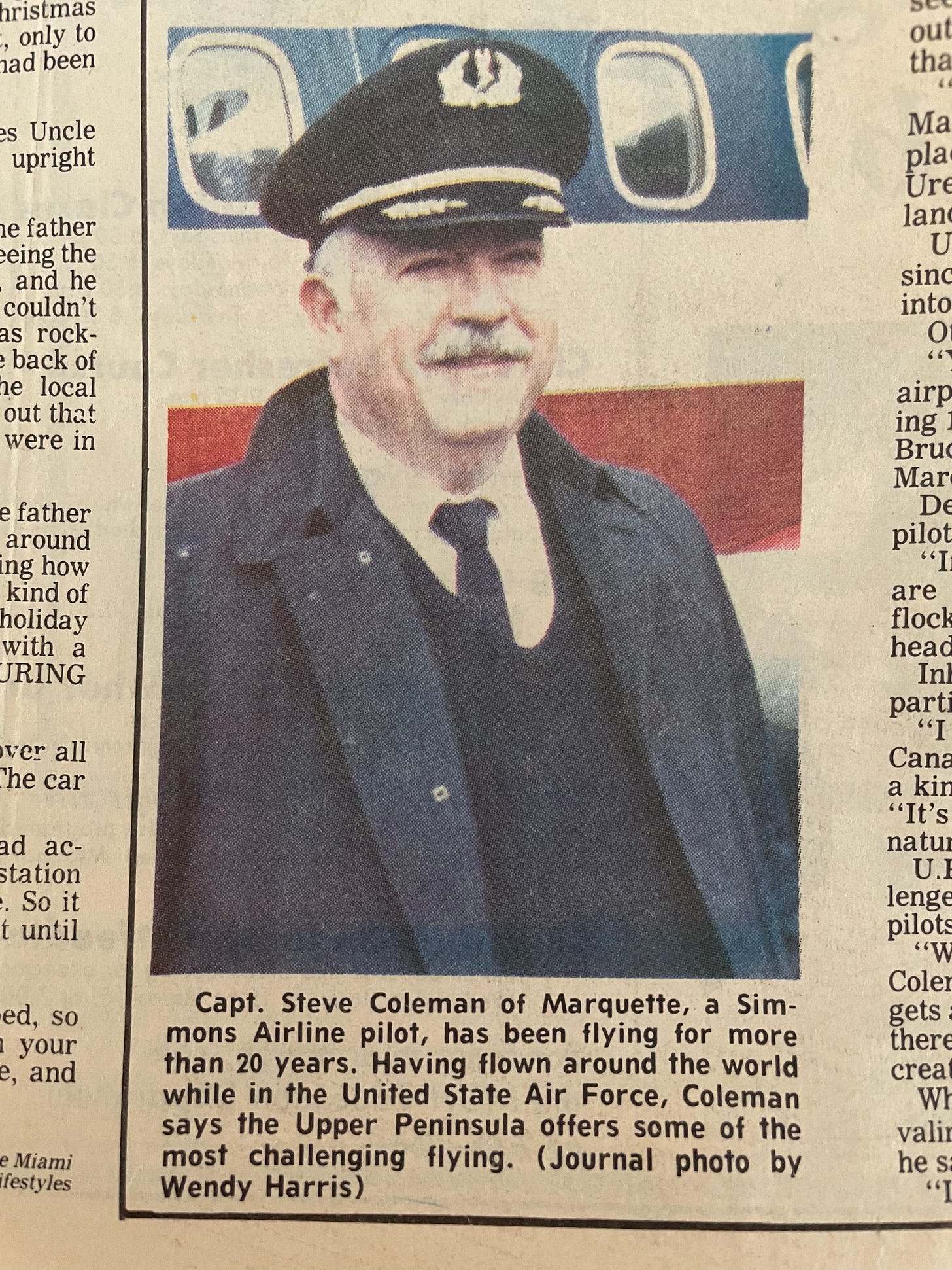 A man with white hair and gray mustache smiles warmly, he is wearing a captain's uniform for a pilot, a trench coat, tie, and sweater vest. He stands in front of a red white and blue passenger plane A man with white hair and gray mustache smiles warmly, he is wearing a captain's uniform for a pilot, a trench coat, tie, and sweater vest. He stands in front of a red white and blue passenger plane