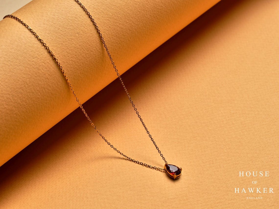 May include: A gold-coloured necklace featuring a teardrop-shaped, dark red gemstone pendant. The fine chain and pendant are set against a warm, orange-toned backdrop. The brand name "HOUSE OF HAWKER ENGLAND" is visible in the lower right corner. May include: A gold-coloured necklace featuring a teardrop-shaped, dark red gemstone pendant. The fine chain and pendant are set against a warm, orange-toned backdrop. The brand name "HOUSE OF HAWKER ENGLAND" is visible in the lower right corner.