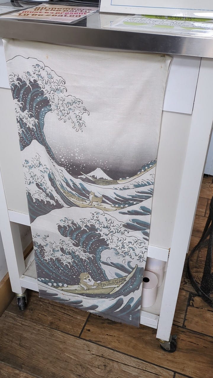 Art in menya hosaki, a take on the famous 'Great Wave' painting of Mount Fuji with Dogs surfing, and a hand-drawn chalkboard sidewalk sign with menya hosaki written on something that looks like a cup of noodles and little faces and characters.
