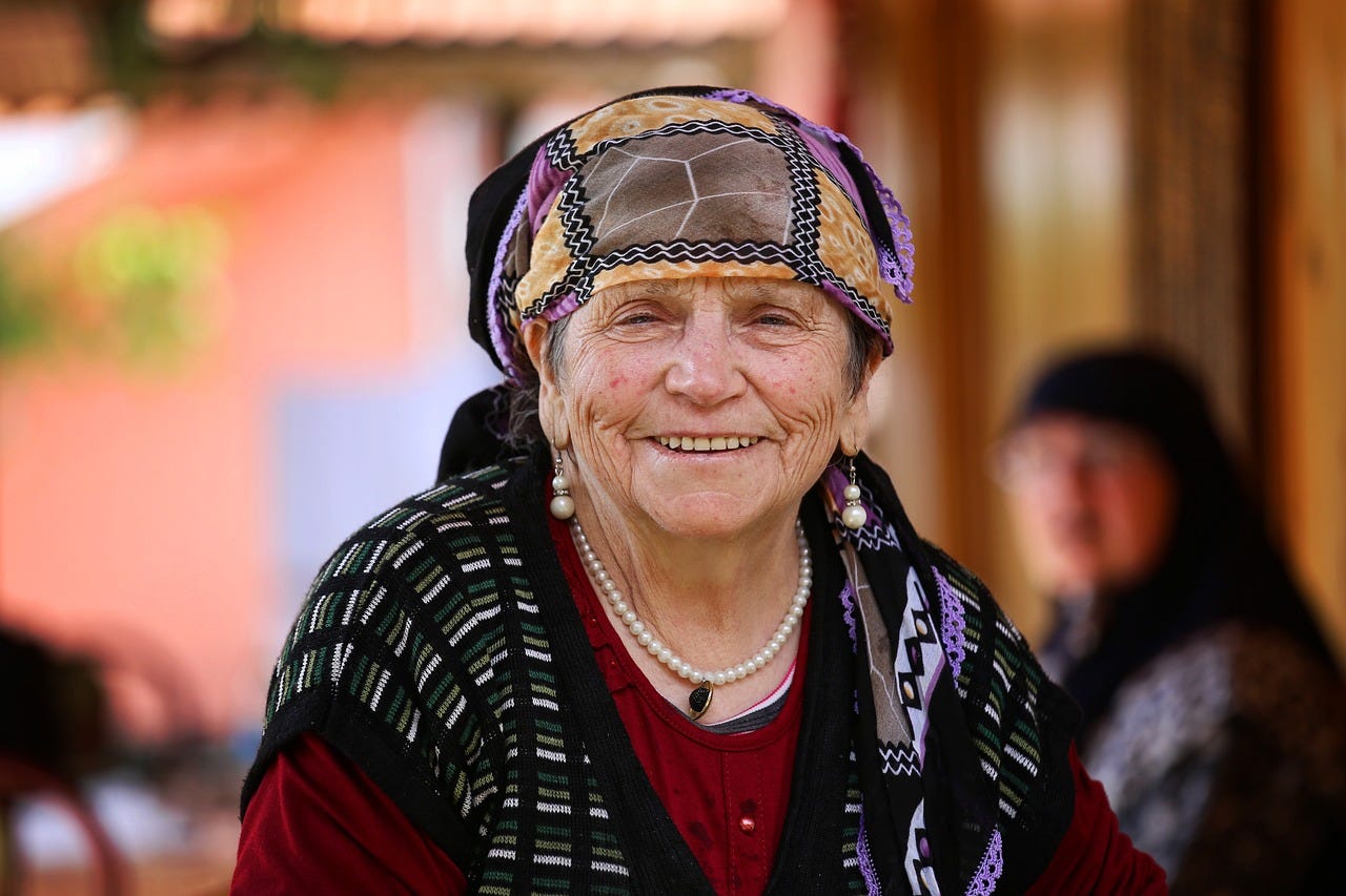 An old woman with a scarf covering her head, wearing pearl earrings and necklace, grins with joy. An old woman with a scarf covering her head, wearing pearl earrings and necklace, grins with joy.