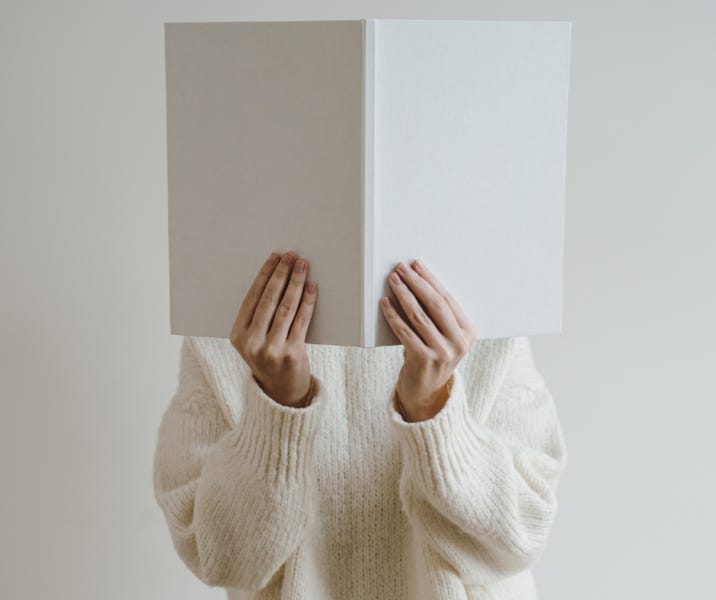 A woman in white against a gray background holds a blank book in front of her face 