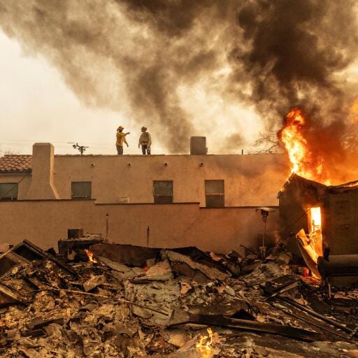 Fire fighters stand on a roof while a building blazes next to them. Rubble remains of a building in the foreground. 