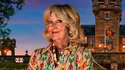A woman in a patterned blouse smiles to camera, standing in front of a castle by night A woman in a patterned blouse smiles to camera, standing in front of a castle by night
