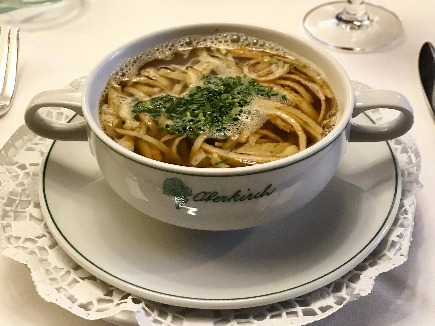A small, double-handled bowl of amber-coloured broth packed with strips of pancakes and garnished with a lot of chopped chives