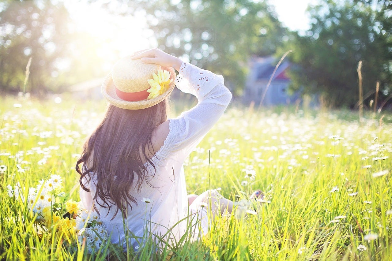 In a sunlit field full of daisies, a woman with long, brown hair sits with her back to you, while resting her hand on her sun hat.