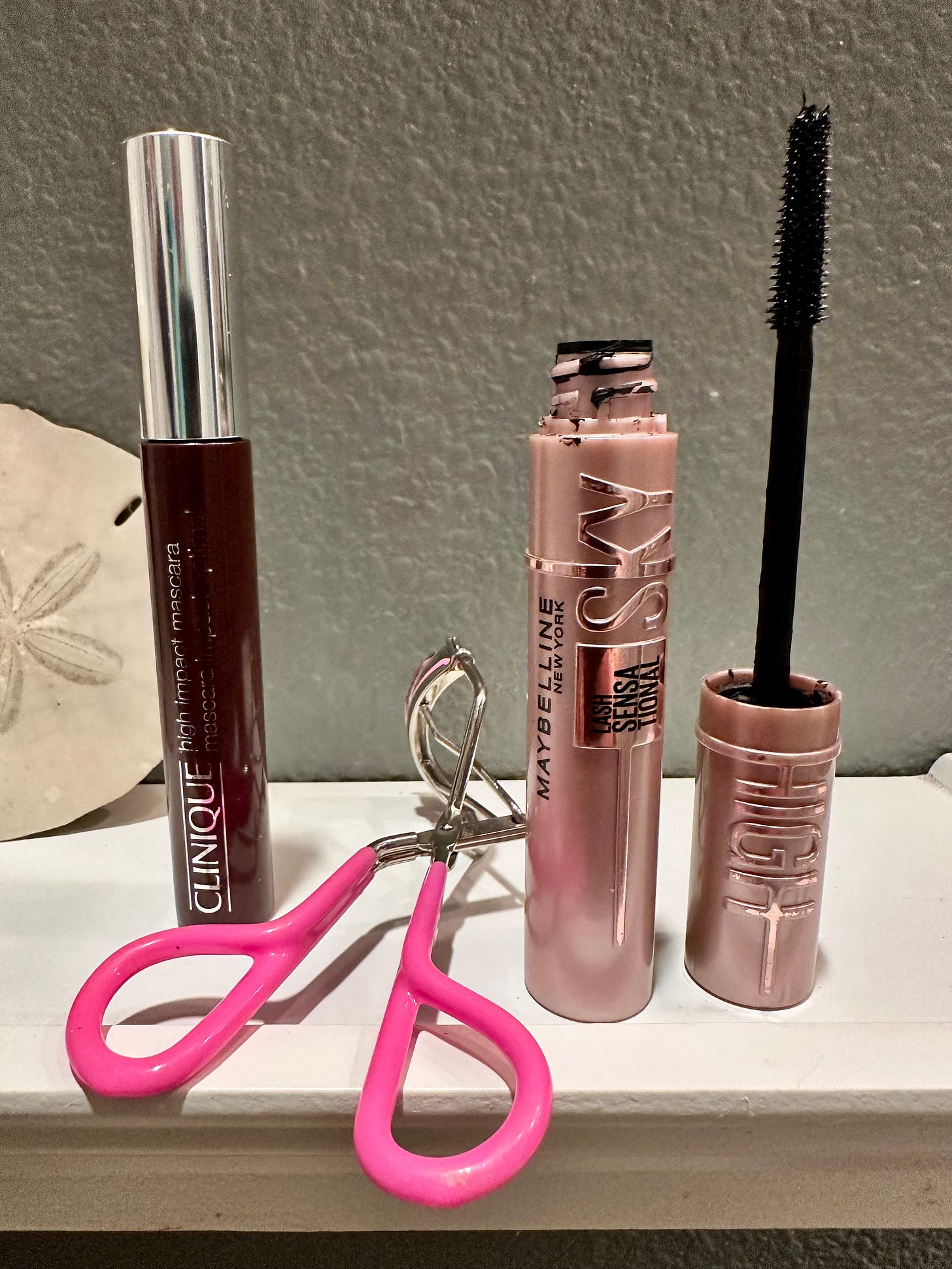 A side-by-side product shot for The Beauty Ed® featuring Maybelline Lash Sensational Sky High Mascara in Black and Clinique High Impact Mascara in Brown. The Maybelline wand is shown unscrewed to reveal the flexible Tower brush, alongside a pink lash curler and the Clinique silver-capped tube. Top-rated mascaras for sensitive eyes as reviewed by THE LAB mentees.