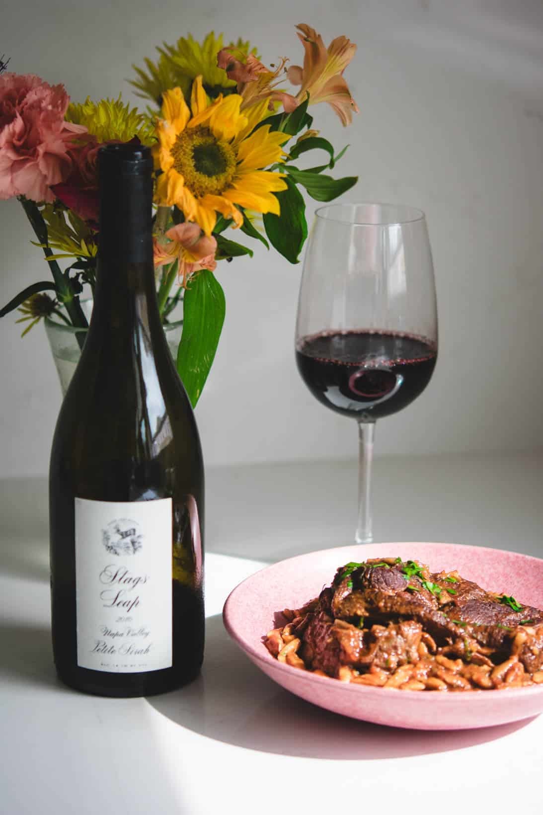 Stags' Leap Petite Sirah wine bottle and glass with flowers in the background and a pink bowl of lamb manastra Stags' Leap Petite Sirah wine bottle and glass with flowers in the background and a pink bowl of lamb manastra