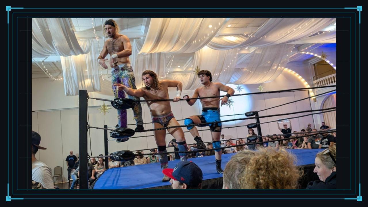 Three professional wrestlers pose along the ropes and corner of a wrestling ring while facing the audience. One stands elevated on the turnbuckle, another points outward, and a third leans against the ropes as spectators watch from close range.