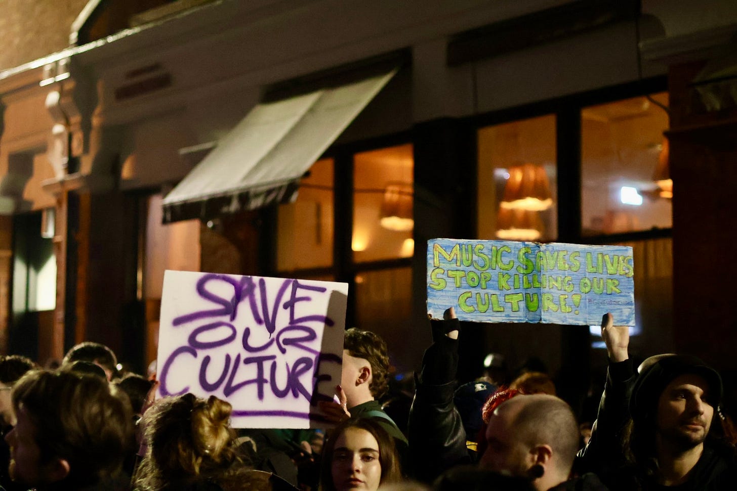 A crowd of protesters gathered outside a lit hotel building at night, holding handmade signs that read “Save Our Culture” and “Music Saves Lives – Stop Killing Our Culture!” as they raise them above their heads. A crowd of protesters gathered outside a lit hotel building at night, holding handmade signs that read “Save Our Culture” and “Music Saves Lives – Stop Killing Our Culture!” as they raise them above their heads.