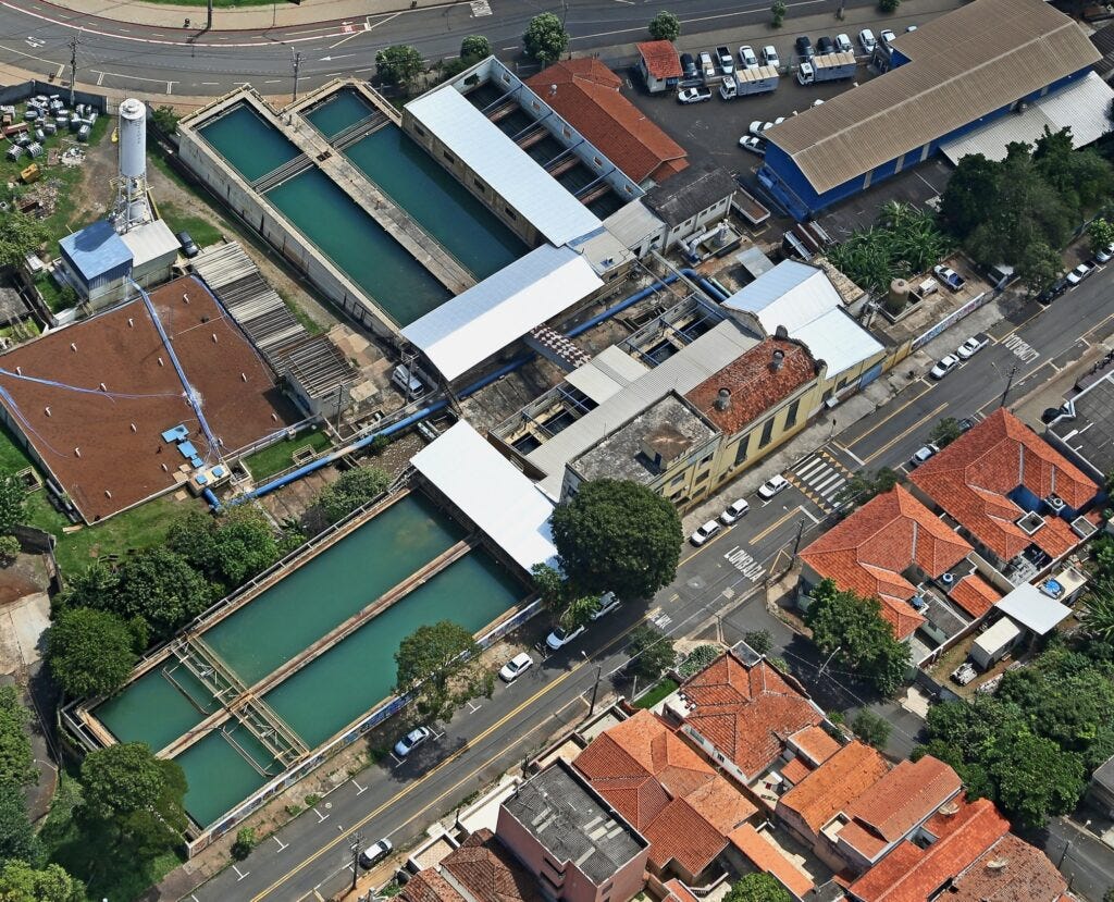 A imagem é uma vista aérea de uma estação de tratamento de água. Vários tanques retangulares com água verde estão visíveis, juntamente com edifícios com telhados vermelhos e brancos. Há também uma estrada com carros estacionados e em movimento, e árvores ao redor da instalação. A estação parece estar localizada em uma área urbana. A imagem é uma vista aérea de uma estação de tratamento de água. Vários tanques retangulares com água verde estão visíveis, juntamente com edifícios com telhados vermelhos e brancos. Há também uma estrada com carros estacionados e em movimento, e árvores ao redor da instalação. A estação parece estar localizada em uma área urbana.