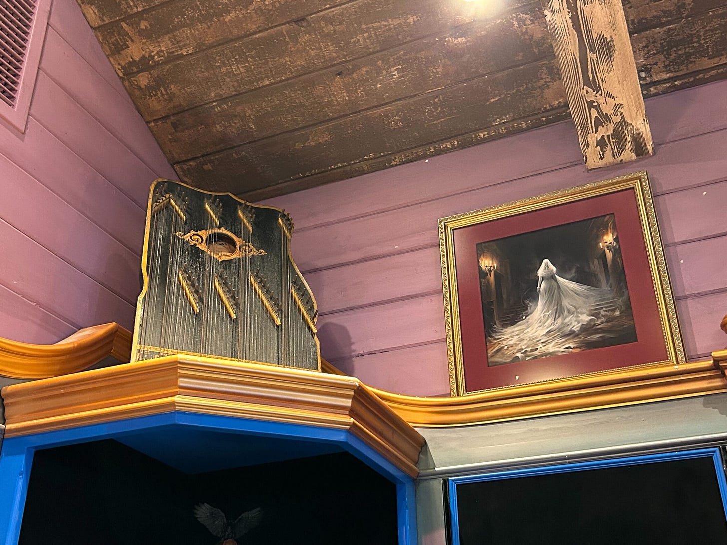 A wooden shelf holds a vintage string instrument and a framed painting of a ghostly figure in a white dress. Echoes of Disneyland's Haunted Mansion linger as the walls and ceiling, painted in purple and brown tones, invite you to Madame Leota's Somewhere Beyond.