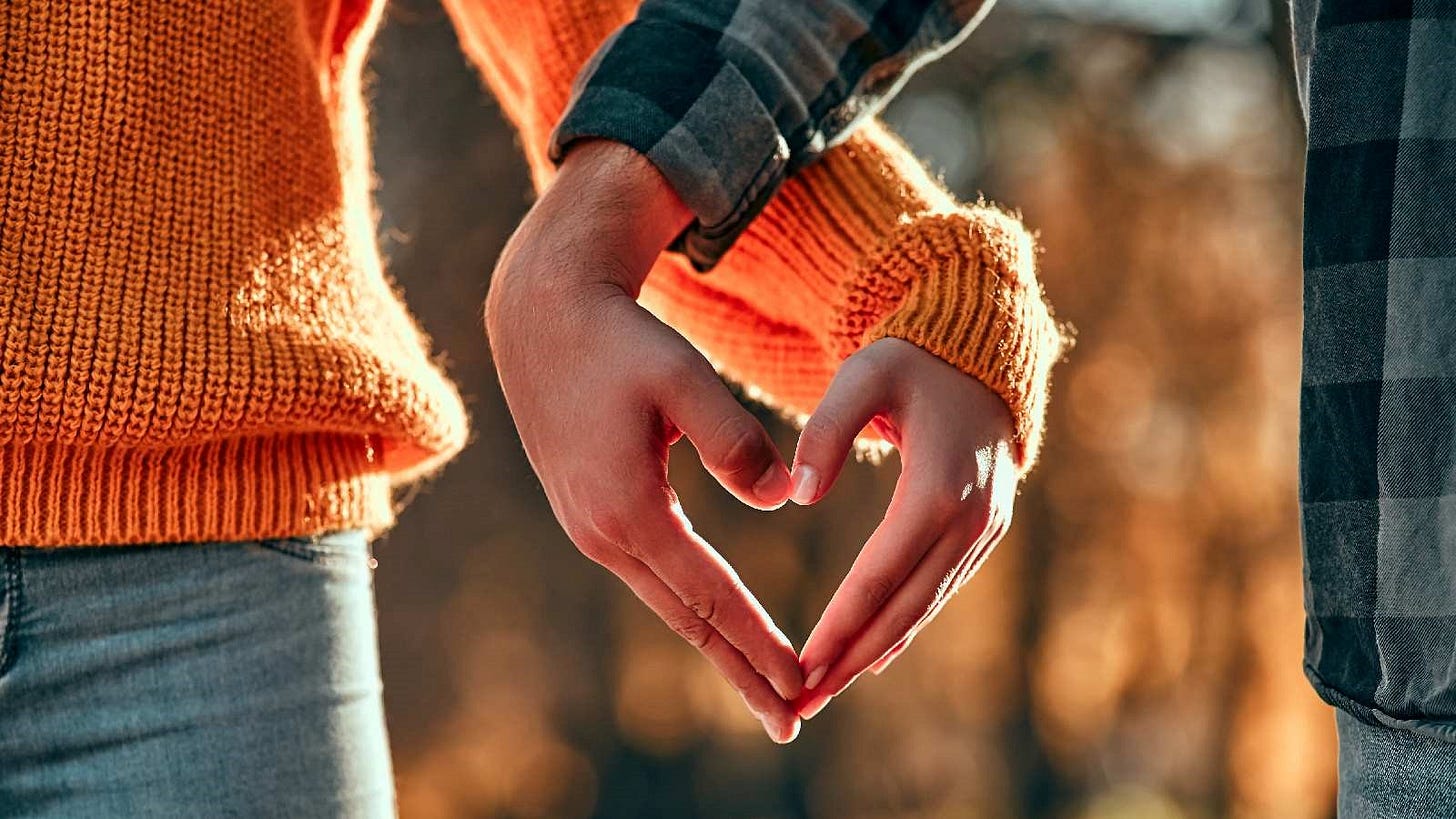 Close-up of two people outdoors wearing cozy sweaters, forming a heart shape with their hands. The warm lighting and autumn colors convey a feeling of affection and connection.