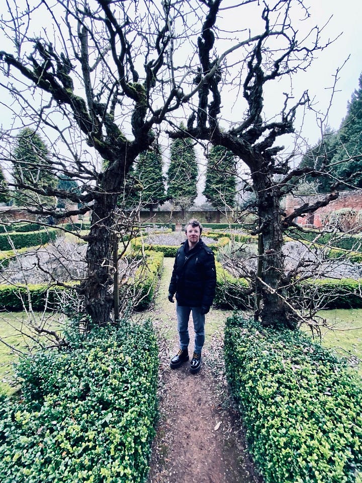 Two photos of me, a white trans masc with round glasses and dark blonde/light brown hair – one in the Abbey gardens, framed by a bare topiary tree archway, the second inside trying on a black top hat