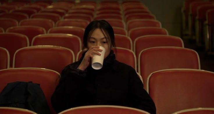 This may contain: a woman sitting in an empty auditorium drinking from a cup