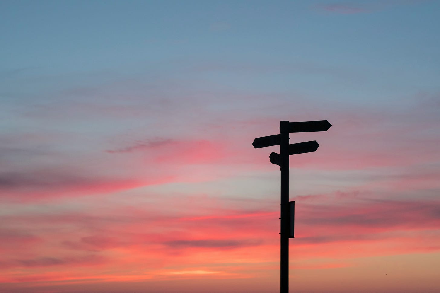 A backlit, multidirectional street sign with a colorful sunset