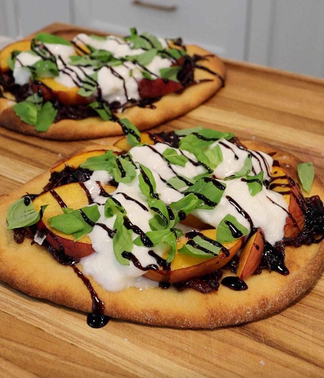 Two peach and burrata flatbreads topped with caramelized red onions, fresh basil, and a drizzle of balsamic glaze on a wooden cutting board.