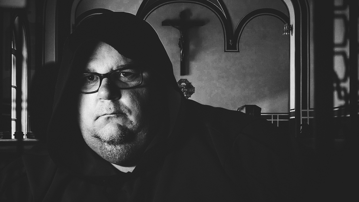 A man in a hooded cloak and glasses sits in a dimly lit church with a crucifix in the background, evoking a contemplative, monastic atmosphere.