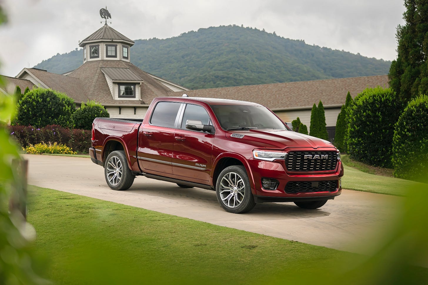 A red 2025 Ram 1500 Tungsten is parked on a driveway in front of a large home tucked into the mountains.