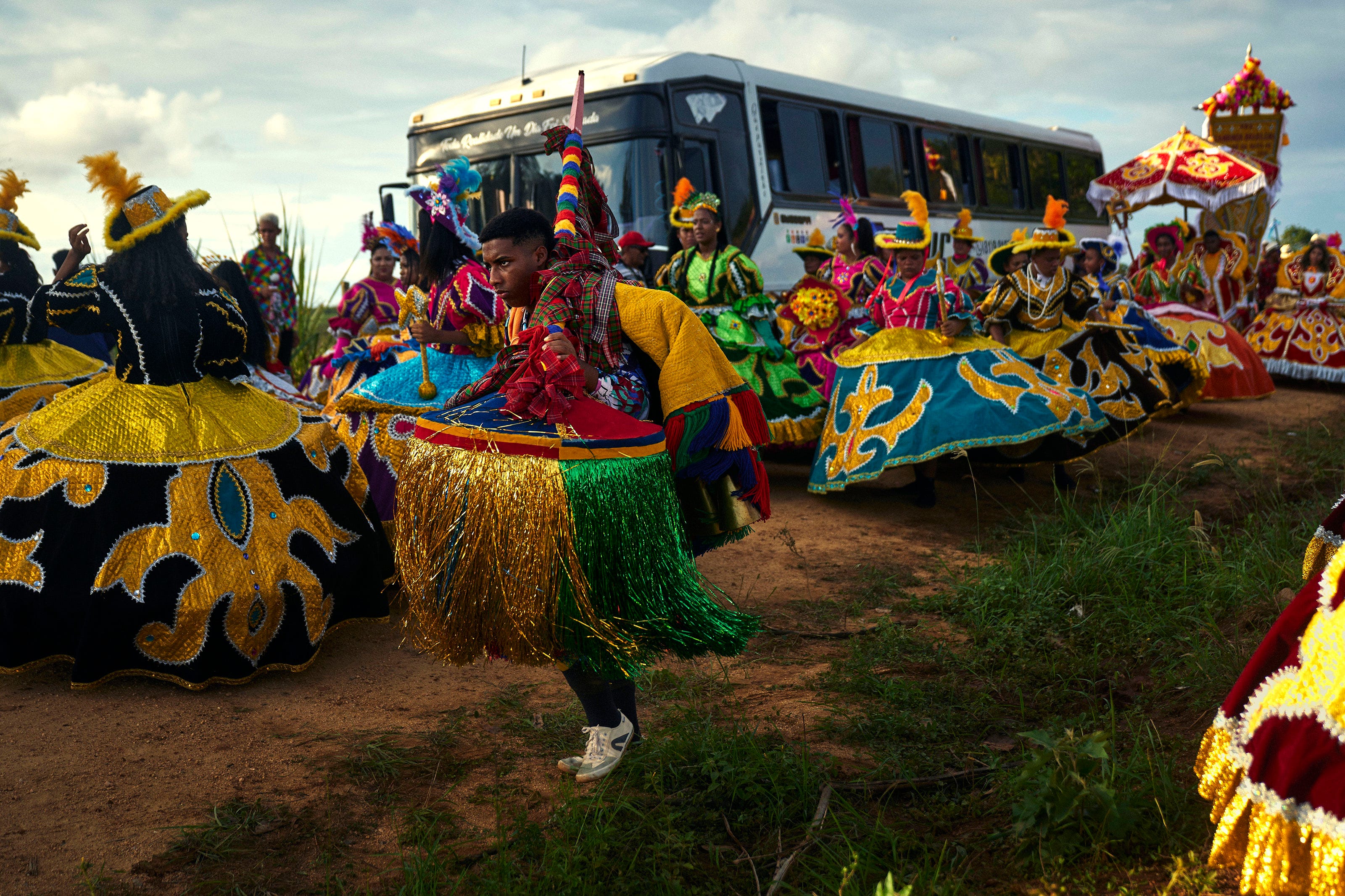 Dancers in colorful, elaborate costumes perform outdoors near a white bus, with green grass and dirt underfoot. Bright fabrics and decorative hats fill the lively scene, suggesting a festival or traditional celebration.