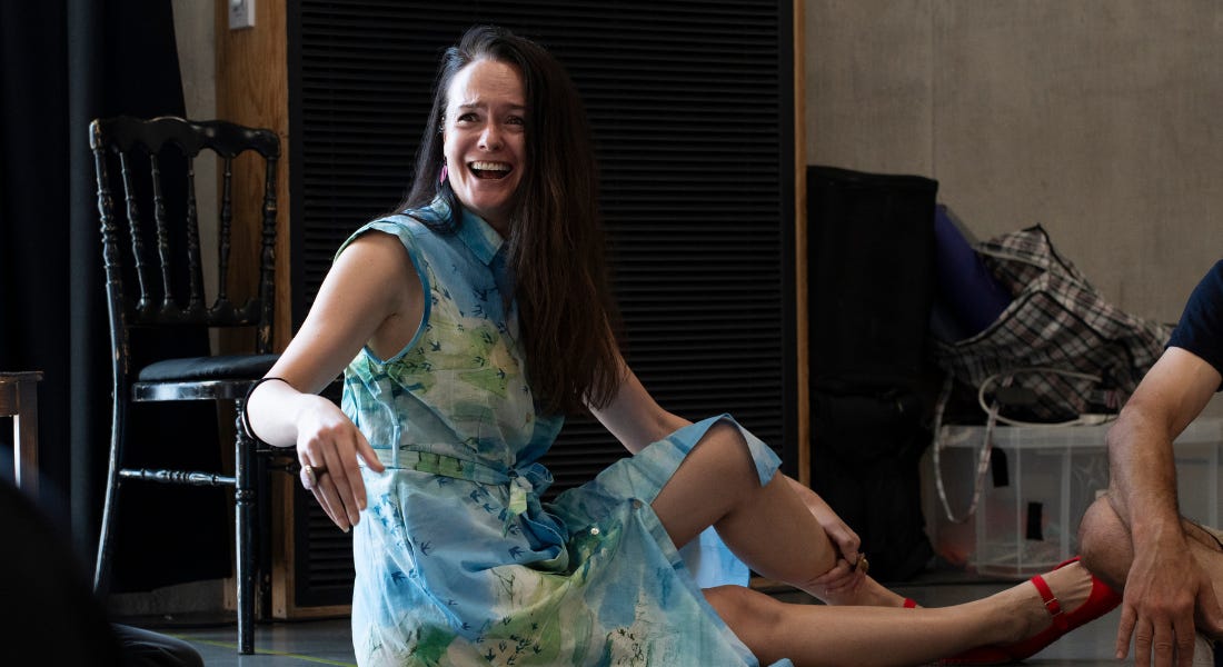 A woman in a blue and green patterned dress with long dark hair sits on a rehearsal room floor, with one leg outstretched. She has one arm slightly raised and is laughing. A woman in a blue and green patterned dress with long dark hair sits on a rehearsal room floor, with one leg outstretched. She has one arm slightly raised and is laughing.