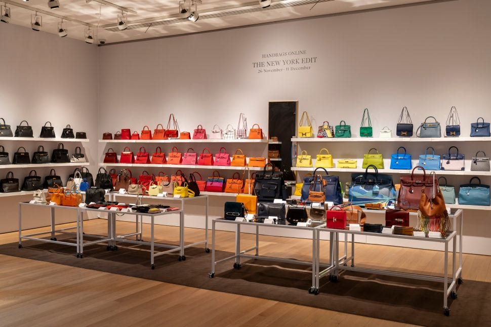 An exhibition view shows rows of colorful luxury handbags displayed on shelves and tables beneath wall text for “Handbags Online: The New York Edit.”