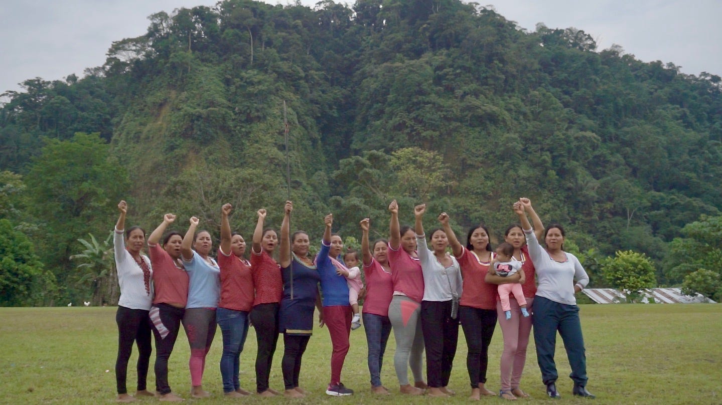 13 members of the Yuturi Warmi stand sideways to the camera with their fists in the air, chanting, on a cloudy day outside in a field