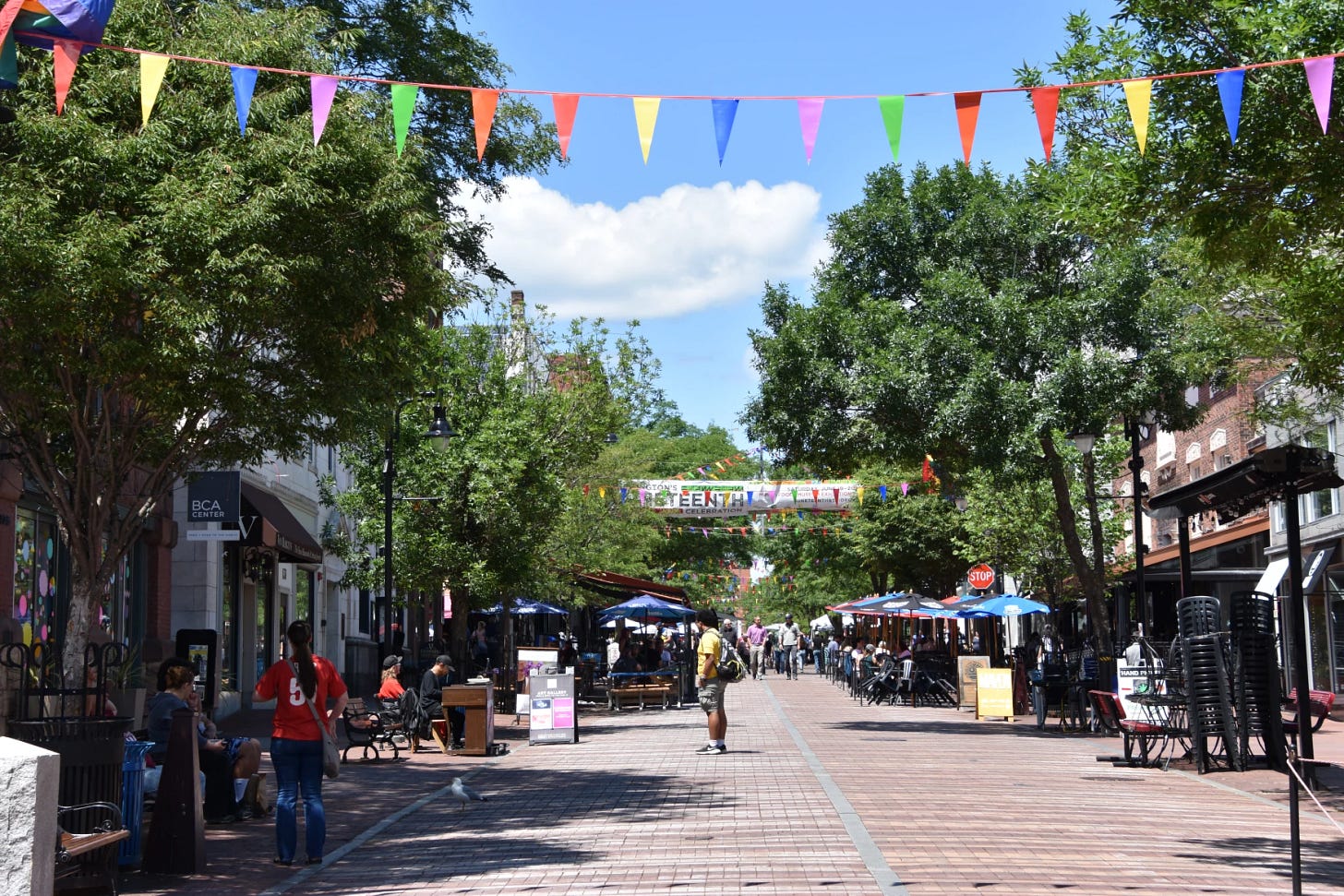 Pedestrians stroll and dine along Burlington’s brick-paved Church Street Marketplace on a sunny day, with colorful flags strung overhead and trees lining the pedestrian mall. Pedestrians stroll and dine along Burlington’s brick-paved Church Street Marketplace on a sunny day, with colorful flags strung overhead and trees lining the pedestrian mall.