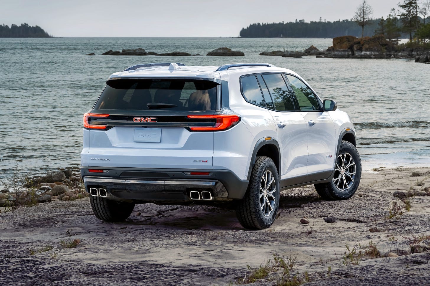 A white 2024 GMC Acadia AT4 is parked on sand near an ocean bay.