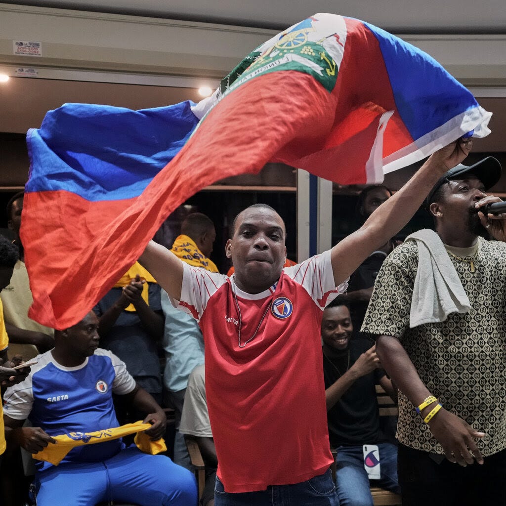 A man in a red and white top waves a multicolor flag as fans in Haiti watch a 2026 FIFA World Cup qualifying match.