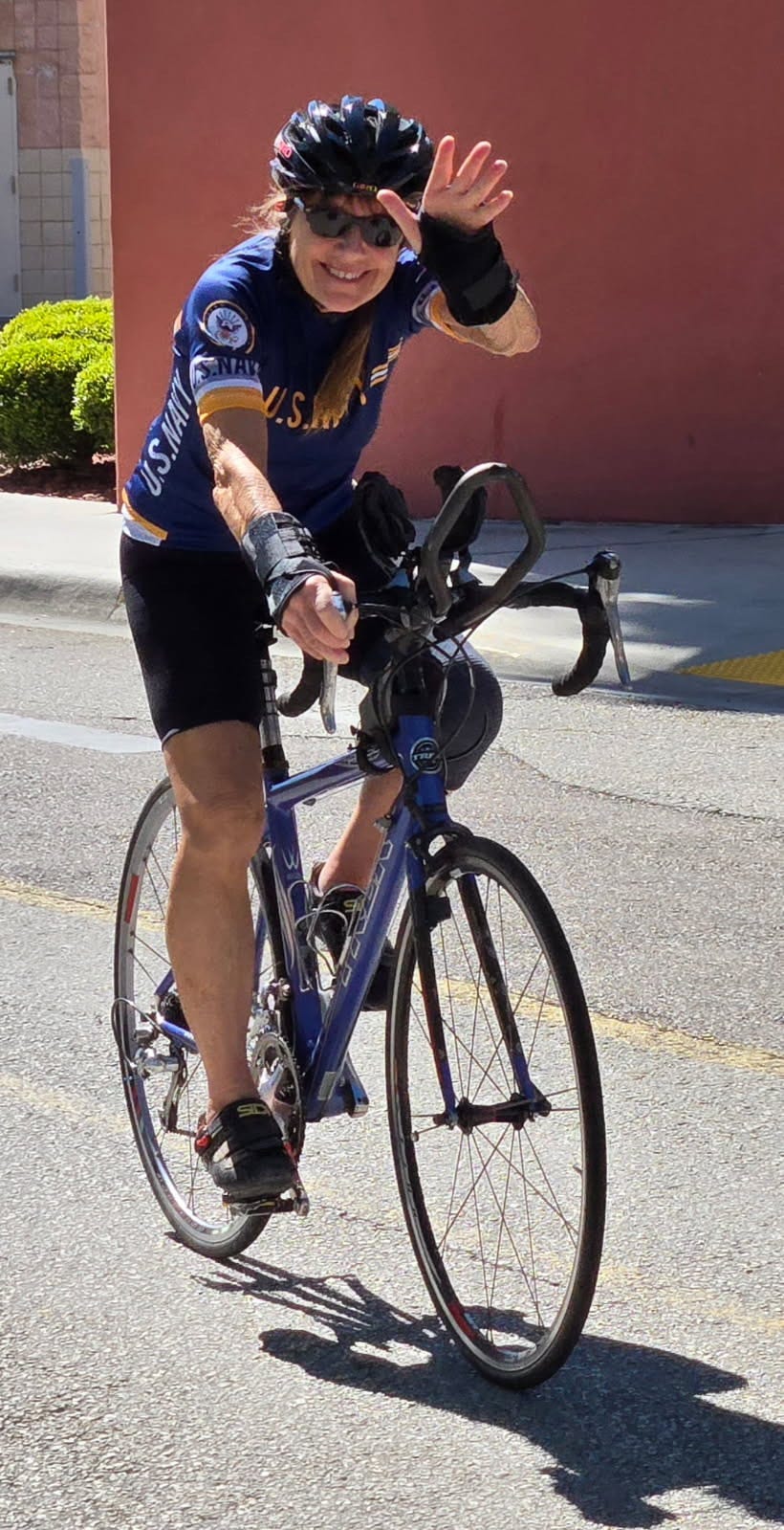 The image shows a person riding a road bike on a city street. The cyclist is smiling, wearing a helmet, sunglasses, and a blue cycling jersey with "U.S. NAVY" and the U.S. Navy emblem clearly visible. The person is waving with one hand while the other hand is on the handlebars. The background includes a red wall, some greenery, and part of a sidewalk. The cyclist is also wearing black shorts, black cycling shoes, and wrist braces. The bike appears to be blue with drop handlebars, and the rider is mid-motion, enjoying the ride. The image shows a person riding a road bike on a city street. The cyclist is smiling, wearing a helmet, sunglasses, and a blue cycling jersey with "U.S. NAVY" and the U.S. Navy emblem clearly visible. The person is waving with one hand while the other hand is on the handlebars. The background includes a red wall, some greenery, and part of a sidewalk. The cyclist is also wearing black shorts, black cycling shoes, and wrist braces. The bike appears to be blue with drop handlebars, and the rider is mid-motion, enjoying the ride.
