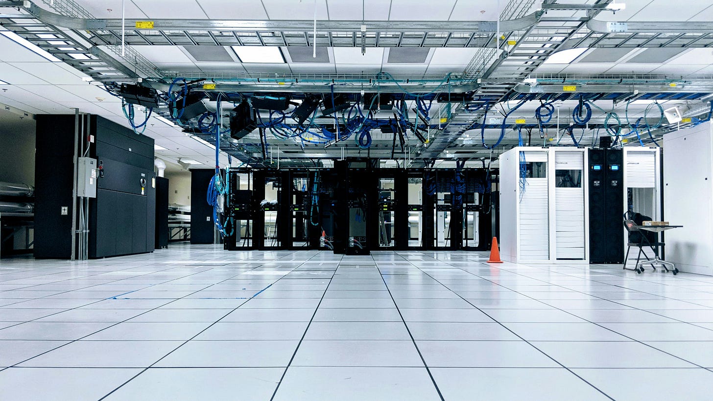 A photo of a sterile white server room with blocks of vertical casing for the servers in black and white with bunches of blue cables hanging above.
