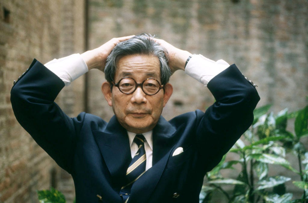 La imagen muestra a un hombre de mediana edad, con gafas y cabello gris, que luce un traje oscuro. Tiene una expresión seria y una postura formal, con las manos levantadas a los lados de su cabeza, como si estuviera frustrado o pensativo. El fondo parece ser un ambiente exterior con vegetación, sugiriendo que la imagen fue tomada en un entorno natural o de jardín.