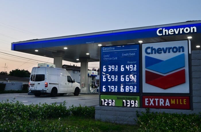 A panel van drives up to a Chevron gas station's pump at dusk. A lit-up sign advertises prices ranging from $6.39 to $7.39 a gallon.