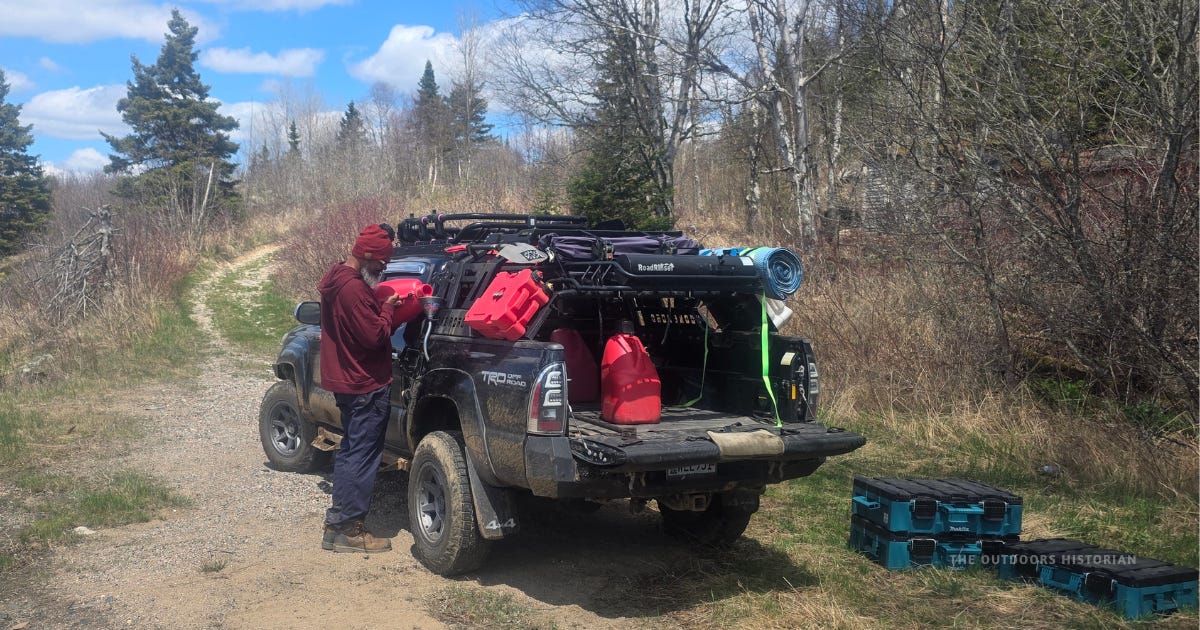 A Toyota Tacoma is parked at the end of a dirt two-track road with the tailgate down. Storage bins are on the ground while a person refuels the truck from a red jerry can under sunny blue skies.