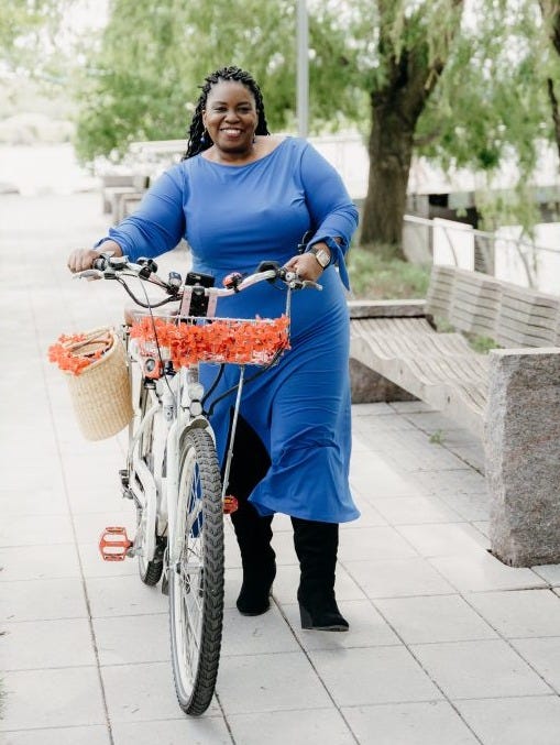 Picture of Renee Moore in a Blue Dress walking her bicycle which has a basket of orange flowers. In the background are trees and benches.