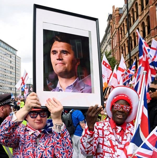 A man in a suit and tie with a Union-Jack pattern and another in a suit with a St. George flag pattern hold up a framed photograph of Mr. Kirk. 