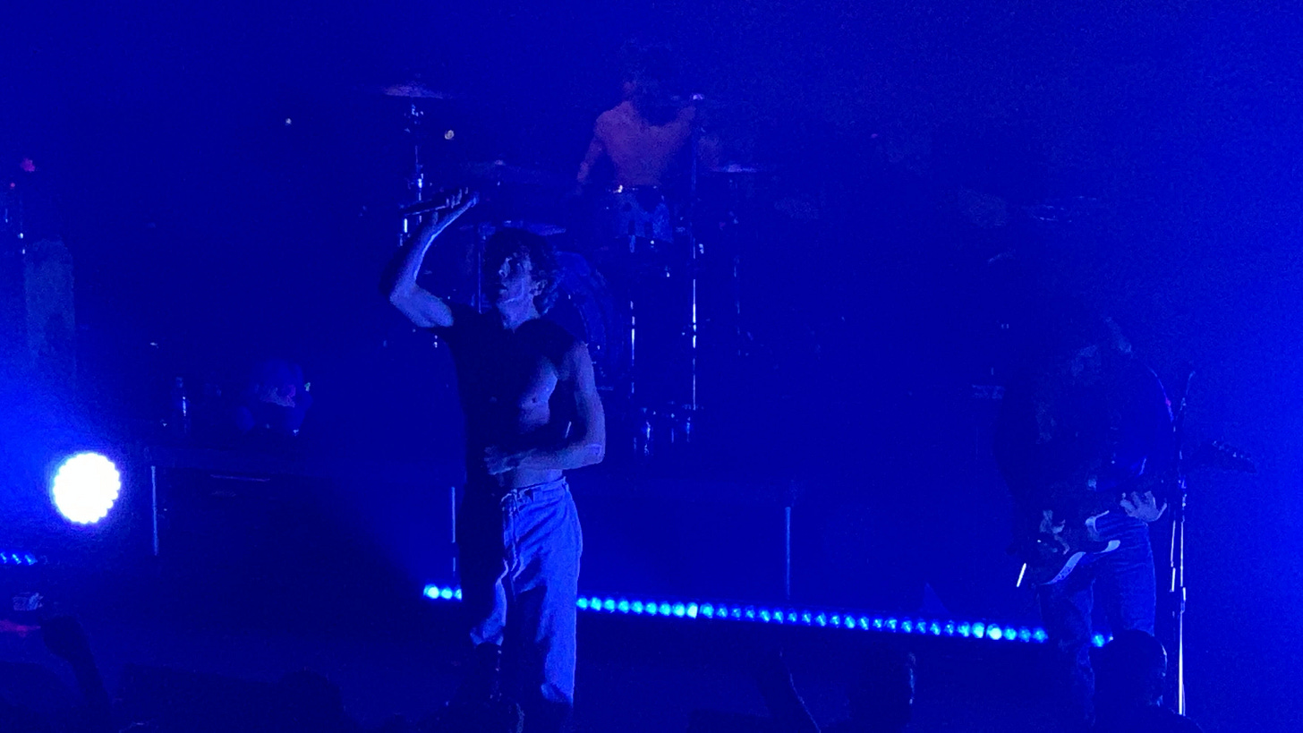 Turnstile at 9:30 Club. Background is deep blue, Brendan is shirtless and has his right arm raised 