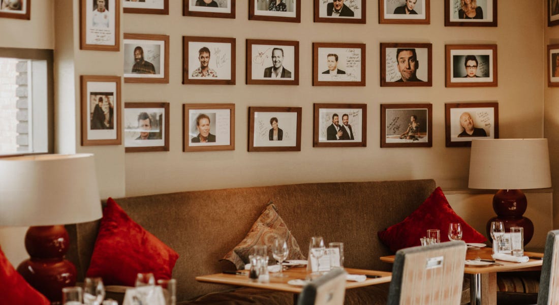 A restaurant with cream curved walls with framed pictures of celebrities, a curved brown couch with red and brown cushions, and wooden dining tables with glasses on top